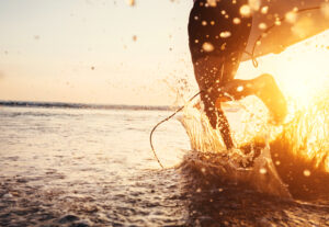 Man surfer run in ocean with surfboard. Closeup image water splashes and legs, sunset light